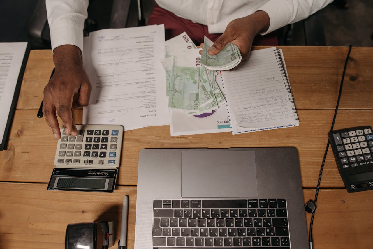 Close-up of a person counting cash with documents and a laptop in an office setting.